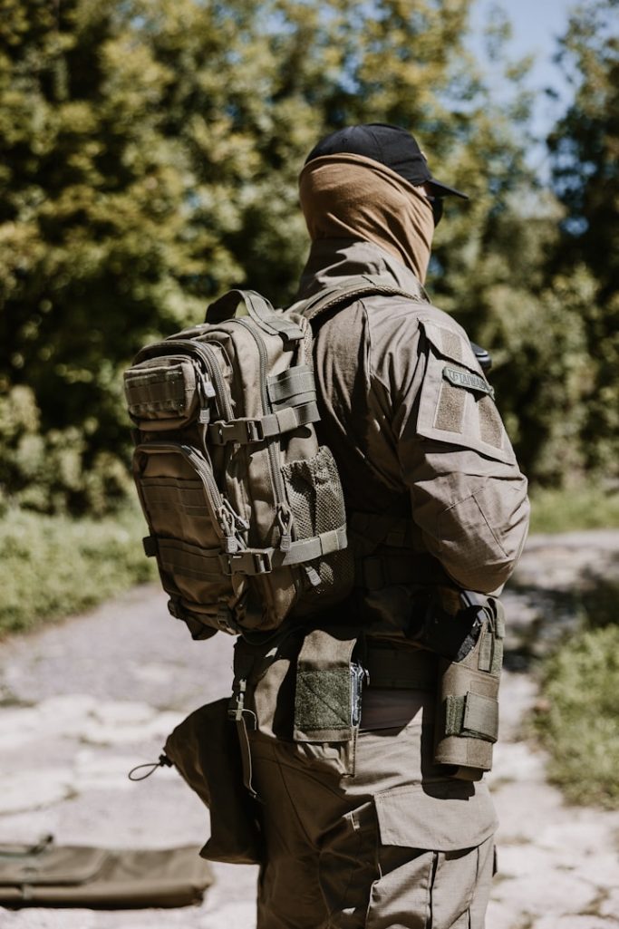 Soldier in tactical gear with a backpack, standing outdoors among trees.